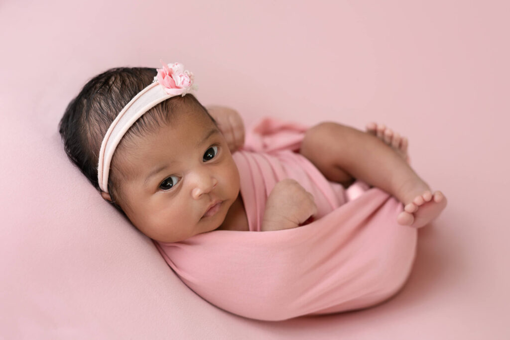 baby girl, awake, looking at camera and wrapped in pink on a pink backdrop