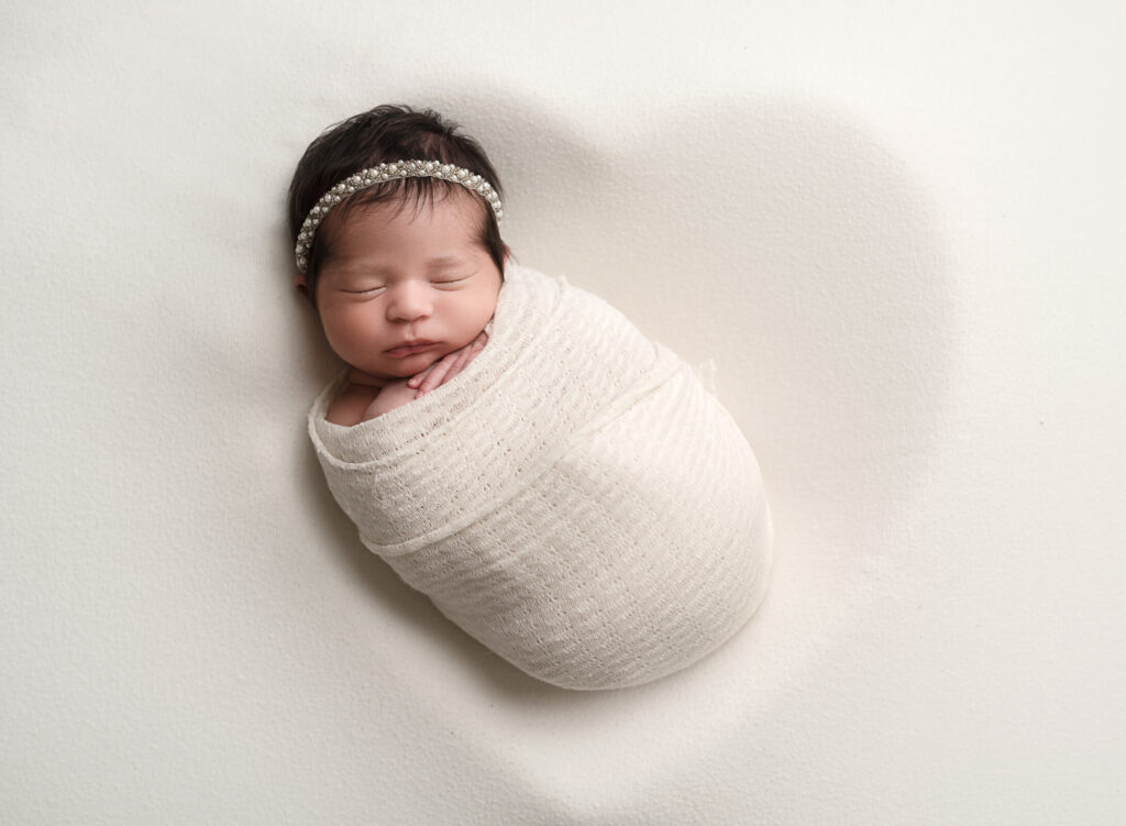 baby girl wrapped in white, and laying on cream backdrop with the outline of a heart